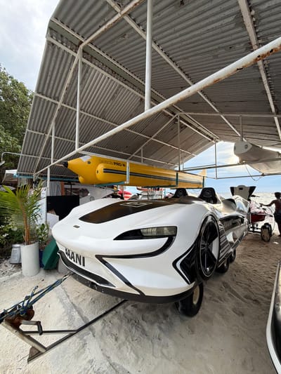 The image depicts an outdoor scene in Maafushi, Maldives, featuring a white and black personal watercraft (PWC) with the name "SAANI" visible on its side. The PWC is positioned on a trailer near a partially covered structure with a corrugated metal roof supported by white painted beams. A yellow "SKIBOB PRO" inflatable water sports device is hanging nearby. A man is vaguely visible in the background near other PWCs on a sandy surface, possibly a beach. A potted palm tree is also visible in the foreground. The lighting is bright, indicating a sunny day.