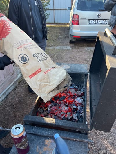 The image depicts an outdoor scene in Заозерное Снт, Russia, centered on a black metal brazier containing glowing red and gray charcoal. A person, identified by a partially visible gloved hand and arm, is pouring birch charcoal from a large brown paper bag labeled "БЕРЕЗОВЫЙ УГОЛЬ" (Birch Charcoal) by "Шефрепс HORECA LINE" into the brazier. An open aluminum beverage can with Cyrillic text "ПИДС" is positioned on the left edge of the brazier's frame, adjacent to a blue and gray handheld torch on the right. In the background, a silver Volkswagen Touran minivan, bearing Russian license plate "У 068 МТ 98 RUS", is parked. Portions of two additional individuals are discernible: one standing to the left of the brazier, and another near the vehicle. The ground consists of dry soil with minimal vegetation, and a light-colored corrugated metal fence is visible behind the vehicle.