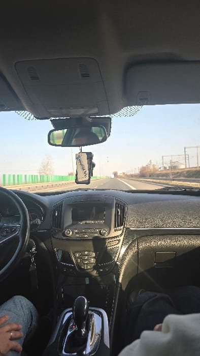 The image displays the interior of a modern vehicle from the front passenger's perspective, looking forward through the windshield, while traveling on a multi-lane highway in the city of Stelnica, Romania.

Inside the vehicle, the driver's left hand is visible on the lower left portion of a dark, multi-functional steering wheel. A gold-colored ring is present on the driver's left ring finger. A set of keys hangs near the steering column. The center console features an automatic gear shifter in the "P" (Park) position. The dashboard is dark, with a central infotainment screen that appears off or showing a dark reflection, flanked by air vents and surrounded by various controls for climate and audio. A white and blue rectangular air freshener hangs from the rearview mirror, which reflects a partial view of the driver's head. The front passenger's right leg, clad in dark trousers, is visible in the lower right foreground, along with a portion of a light-colored garment. Direct sunlight casts bright reflections on the dashboard and center console.

Outside the vehicle, a wide asphalt highway extends into the distance under a clear, bright blue sky. A green metal guardrail runs along the left side of the highway. Sparse, bare trees and dry, brown vegetation are visible alongside the road. In the far distance on the right, several light-colored industrial structures, possibly silos or grain elevators, are observable, accompanied by what appears to be railway lines or power lines. At least one other vehicle is visible in the distance, traveling in the same direction.