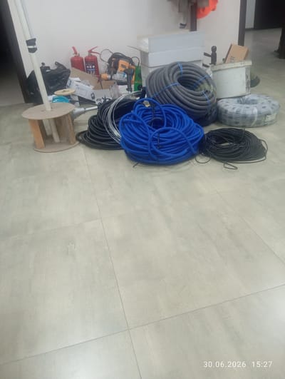 This media file depicts a storage area, likely a workshop or construction site, in Ohangaron, Uzbekistan. The scene is filled with various electrical and construction materials. Prominently featured are large coils of gray and blue corrugated tubing, as well as spools of black electrical wire. There are also two red fire extinguishers, a power drill, and several cardboard boxes. A wooden spool stand is visible on the left side of the image. The floor is tiled with light-colored square tiles. The lighting suggests an indoor environment, likely during daytime. The overall impression is one of organized clutter, typical of a space where work is either in progress or materials are being stored. The timestamp "30.06.2026 15:27" is visible in the bottom right corner, indicating the date and time the image was captured. No people are visible in the frame.