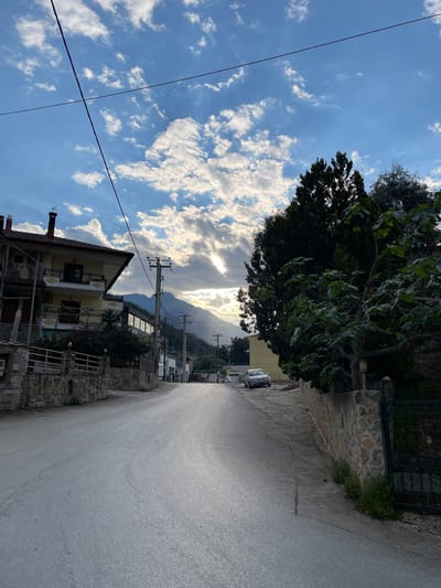 The image depicts a view along a paved road in Skala Potamias, Greece. The road stretches into the distance, where mountains are visible. The sky is blue with numerous white clouds. Power lines are visible across the sky. A large tree overhangs the road on the right side. Buildings and stone walls are situated along the sides of the road. Parked cars are visible in the distance. A two-story building with a chimney is on the left side of the road.