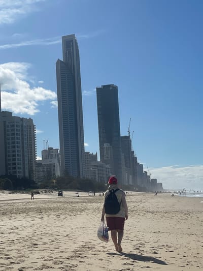 A lone figure, walking away from the camera, traverses a sandy beach in Surfers Paradise, Australia. The individual is wearing a red baseball cap, a light-colored hooded jacket, and maroon shorts. They are barefoot and carrying a white plastic shopping bag in their left hand. In the background, a cityscape rises, dominated by numerous tall, modern buildings. Some of these skyscrapers are under construction, indicated by visible cranes. The beach itself is wide and populated with a few other individuals and beach umbrellas in the distance. The ocean is visible on the right side of the image, meeting the shore with gentle waves. The sky above is a clear blue with scattered white clouds.