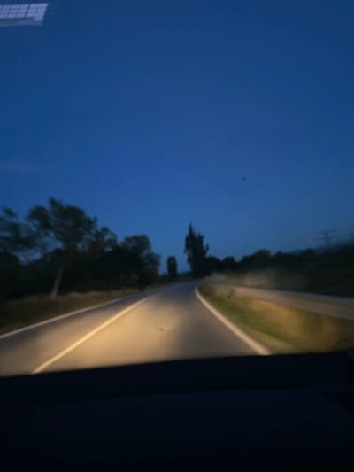 The image shows a view from the perspective of a driver at dusk, looking out at a dark rural road. The sky is a deep blue, transitioning from twilight to night. The headlights of the car illuminate the asphalt road, which has a white line marking the center. Trees and foliage line the sides of the road, appearing as dark silhouettes against the sky. The overall impression is one of a quiet drive through the countryside as darkness descends. It is not possible to determine the specific location within the city of Garmen, Bulgaria, from this image alone.