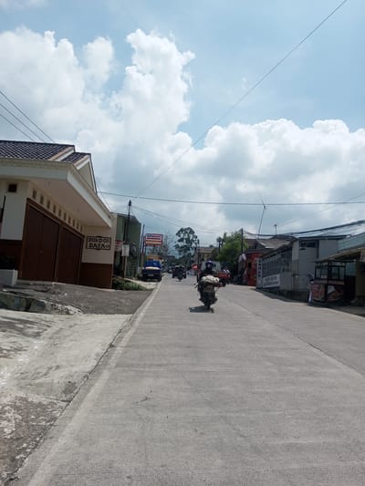 The image depicts a street scene in what appears to be a residential area. A concrete road extends into the distance, with a motorcycle traveling along it. Buildings line both sides of the street, including a structure with brown doors labeled "PONDOK DAYA". Across the street, a sign for "Alfamart" is visible. Power lines stretch across the sky, which is filled with white clouds. Several other vehicles and pedestrians are present in the distance, suggesting a populated area. The photograph’s details are consistent with the description of Zeyathiri District, Myanmar, as a residential district.