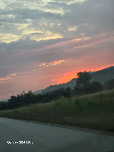 The image displays a rural landscape at either dawn or dusk, captured from what appears to be a moving vehicle, in Gorna Rositsa, Bulgaria. The upper two-thirds of the frame are dominated by a cloudy sky, transitioning in color. The clouds at the top are pale gray and light yellow, gradually becoming darker gray, then soft pink, and finally a vivid orange and red near the horizon. This intense coloration indicates the presence of a rising or setting sun, which is obscured by the distant, dark, undulating silhouettes of forested hills or mountains.
In the midground, closer hills are densely covered with dark green trees, silhouetted against the bright orange sky. A prominent, full tree stands slightly right of the frame's center, its foliage starkly outlined by the intense light. The foreground, occupying the lower third of the image, features a dark gray paved road that angles from the lower-left to the lower-right. A faint lighter line, possibly a shoulder marking, is visible along the left edge of the road. Adjacent to the right side of the road, a strip of tall, brownish-green grass or weeds runs parallel, backed by a gentle slope of shorter, greener vegetation. Minor horizontal motion blur is observable in the foreground elements, consistent with a photograph taken from a moving platform. The text "Galaxy S24 Ultra" is watermarked in white in the lower-left corner. No people, animals, or man-made structures other than the road are visible.