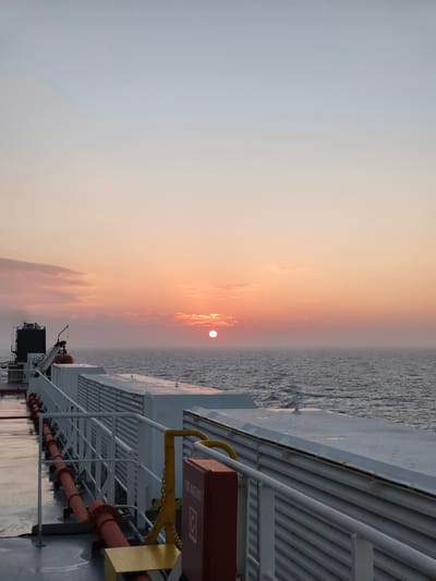 The image depicts a wide-angle view of a ferry deck at sunset. The sky is a gradient of soft blues and oranges, with the sun a bright, round disk low on the horizon, partially obscured by wispy clouds. The ocean stretches out to meet the sky, with gentle waves reflecting the twilight colors.
In the foreground, the deck of the ferry is visible, with a white railing running along the edge. Red pipes are mounted along the deck, and a yellow fire hose box with a red lid is present. The deck appears wet, suggesting recent rain or sea spray. The ferry itself is large and white, with several structures and vents visible. The overall impression is one of calm and vastness, with the setting sun casting a serene glow over the scene. There are no people visible in the image.
The location is not specifically identifiable as Changwon-si, South Korea, as the image provides no geographical markers.