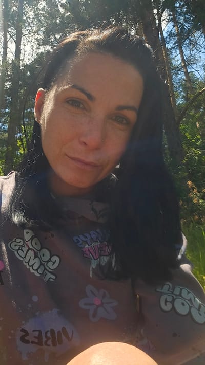 A medium-shot, selfie-style photograph features a woman with dark hair and fair skin outdoors, likely in the city of Dolni Pasarel, Bulgaria. Sunlight filters through trees, casting shadows on her face. She is wearing a grey sweatshirt with colorful graphics, including the words "GOOD TIMES" and "VIBES." The background consists of trees and foliage, suggesting a natural setting. The woman's expression is neutral to slightly smiling, and she appears to be looking directly at the camera.