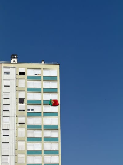 The image shows a tall, light-colored apartment building against a clear blue sky. The building has multiple floors with numerous windows, many of which are covered by white blinds. A Portuguese flag is hanging from a window on the right side of the building. On the roof of the building, there are two chimney structures. The building appears to be situated in an urban environment, likely a residential area, in Charnay-lès-Mâcon, France, given the context provided. The scene is static, with no people or animals visible, and the focus is on the architecture and the flag.
