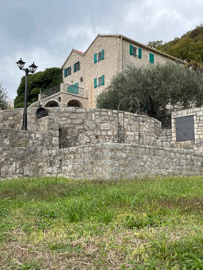 An outdoor scene in Đenjaši Česminovo, Montenegro, depicts a multi-story, light-colored stone building with a reddish-brown tiled roof situated on a hillside under an overcast sky. The building features multiple rectangular windows fitted with bright green wooden shutters; some are open, others are closed. A small balcony with a dark metal railing extends from the front of the house, supported by two stone arches.

In the midground, a substantial terraced stone wall, constructed from rough-hewn grey stones, occupies the lower and central portion of the image. A black, ornate streetlamp with two light fixtures stands on the upper left section of this wall. To its right, a dark, tire-like object rests on the stone surface. Further right, a black metal wellhead or decorative arch structure stands on another section of the stone wall, featuring a small bucket or pulley mechanism. To the far right, a series of stone steps are integrated into the wall, leading up to a dark grey metal door or panel set flush into the stone masonry.

A large olive tree with dense, silvery-green foliage is positioned between the stone wall and the building, partially obscuring the house. Denser green deciduous and evergreen trees cover the hillside behind and to the right of the house. The immediate foreground consists of unkempt grass, displaying varying shades of green and some drier, browner patches. No individuals, animals, or active interactions are present in the scene.