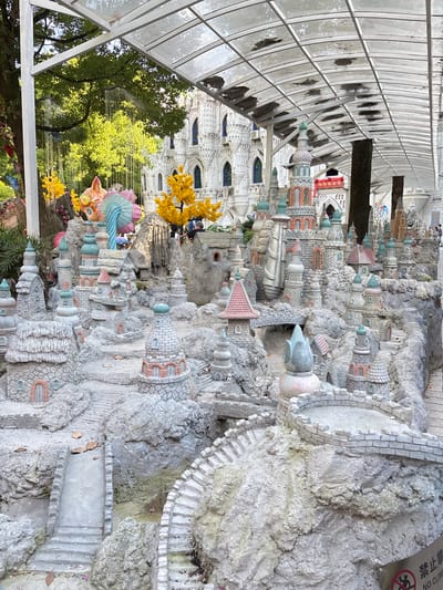 A detailed miniature city made of grey-white stone is under a translucent roof. The miniature city includes castle-like structures, domed towers, and small buildings with brick-like textures. Some structures are adorned with pastel-colored roofs and domes, and others are connected by miniature stone bridges. The surrounding environment has trees, yellow floral decorations, and various colorful ornaments. People can be seen interacting with the exhibit. There is a sign in the upper right corner in Chinese with a symbol that says, "No Climbing" on the structure. It is located in Baqiao, China.
