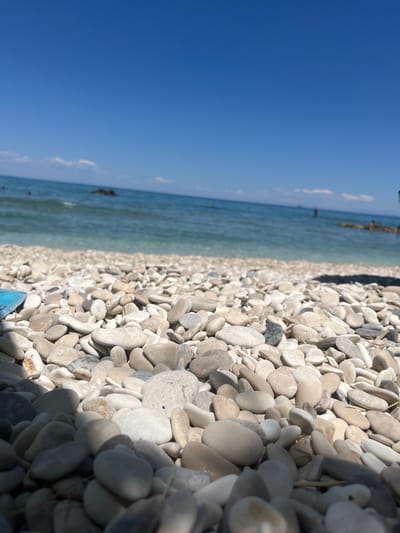 A low-angle, close-up shot shows a pebble beach in Elaties Municipal Unit, Greece, with the clear blue sea and sky in the background. The foreground is dominated by smooth, varied-colored pebbles. In the middle ground, the sea is a turquoise hue, with gentle waves lapping the shore. A few dark rocks are visible in the water, and some indistinct figures of people can be seen swimming or wading in the distance. The sky is a vibrant, cloudless blue. The lighting suggests a sunny day.