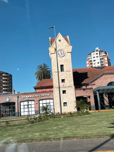 The image shows a Starbucks Coffee location in Tigre, Argentina, next to a clock tower. The Starbucks is a brick building with white lettering on its facade. A tall palm tree grows next to the building. The clock tower is made of light-colored stone and has a clock face on its upper portion. The sky is clear blue. There is a grassy area between the building and a road. A few other buildings are visible in the background.