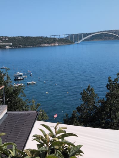 The image depicts a serene coastal scene in Kraljevica, Croatia, with a prominent bridge spanning across the blue water in the background. Several boats, including a jet ski, are anchored in the bay, with buoys marking their positions. Lush green trees frame the foreground, and part of a tiled roof and white awning are visible, suggesting a residential or hospitality setting. The sky is clear and blue, indicating a sunny day. The Krk Bridge, a significant landmark connecting the island of Krk to the mainland, dominates the horizon. The water appears calm, reflecting the clear sky.