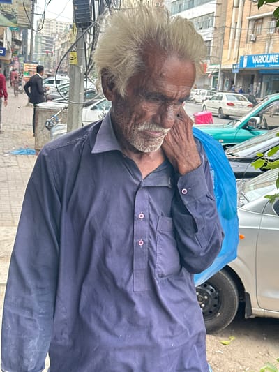 The media file is a portrait-oriented photograph of an elderly man with light-colored hair and a beard. He is wearing a dark blue collared shirt with pink buttons. His right hand is raised to his mouth, and he appears to be speaking or in thought. Behind him, a blue plastic sheet is visible, and further back, a light-colored vehicle is parked. The background suggests an urban street scene with buildings, wires, and other vehicles. In the lower part of the image, other people are visible in the distance, as well as what appears to be a street market or shops. The overall lighting suggests it is daytime. A sign with the word "VARIOLINE" in blue and white lettering is visible on a building in the background, and another sign with yellow text on a pole. The setting appears to be a busy street in Karachi Division, Pakistan.