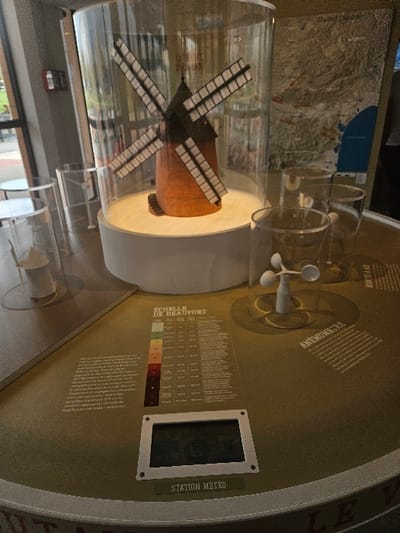 The scene depicts an interior exhibition space, likely a visitor center, in Avignonet-Lauragais, France, highly probable to be associated with the Moulin à vent de Cugarel. A detailed scale model of a traditional European windmill, featuring an orange-brown body, a dark conical roof, and four sets of sails with white segments, is centrally displayed within a clear cylindrical acrylic enclosure on an illuminated white circular pedestal.
This exhibit is situated on a large, olive-green circular table. To the right of the windmill model, a white three-cup anemometer model is visible within another clear cylindrical container, labeled "ANEMOMETRE" in French. Adjacent to this, a rectangular graphic displays the "ÉCHELLE DE BEAUFORT" (Beaufort Scale) with a vertical color gradient and corresponding text descriptions. Below this graphic, an embedded digital screen labeled "STATION MÉTÉO" (Weather Station) presents what appears to be weather data, including a small compass rose and numerical values. Several other transparent cylindrical display cases of varying sizes are positioned on the table.
The background features large glass windows, revealing a paved outdoor area and ambient light. A red fire alarm panel is mounted on a pillar to the left. A large topographical or thematic map is partially visible on a wall behind the main exhibit. No individuals are present within the frame.