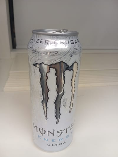 A silver can of Monster Energy "Ultra" is placed on a white surface. The can is upright and slightly angled towards the viewer, showing the brand's distinctive claw mark logo in a darker shade of silver. The text "ZERO SUGAR" is visible near the top of the can, and "MONSTER ENERGY ULTRA" is printed towards the bottom. The background is out of focus but appears to be a neutral-colored wall and shelving. The lighting is soft and diffused. This scene is not located in the city of Anderlecht, Belgium.