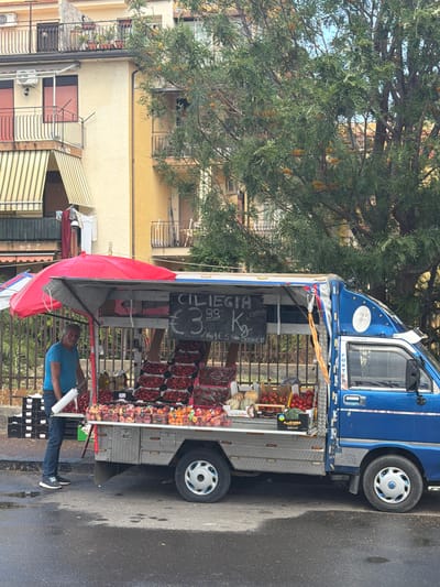 The image shows a mobile fruit and vegetable stand, operated from the back of a blue truck, parked on a wet street in what appears to be a residential area. The stand is laden with cherries and peaches, with a handwritten sign advertising "CILIEGIA" (cherries) at €3.99 per kilogram and another price of €5 for a unit. A man in a blue shirt is visible, possibly the vendor, interacting with customers. The scene is in Chianchitta, Italy, indicated by the language on the sign. In the background is a yellow building with balconies and a tree. The Super Porter brand of the truck is noted.