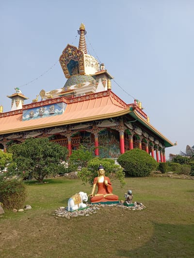 The image depicts a large, ornate Buddhist temple or monastery in Lumbini Sanskritik, Nepal. The structure is multi-tiered, with a prominent stupa-like tower adorned with colorful patterns and gold accents at the top. The main building has a wide, overhanging orange roof supported by numerous red pillars with intricate designs. The walls feature murals and decorative elements.
In the foreground, there is a manicured lawn with several bushes and trees. On the lawn, a seated Buddha statue is present, colored orange and gold. To the left of the Buddha statue, there's a white elephant statue, and to the right, a small grey statue of a meditating person. The statues are situated on a bed of light-colored stones.
The sky is clear and light blue. The overall impression is that of a well-maintained religious site.