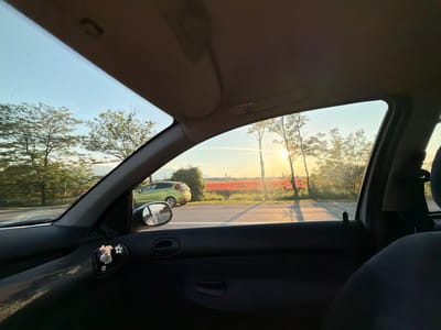 The photograph is taken from inside a car, looking out through the front passenger window and the windshield. The viewpoint is low, capturing the interior of the car including the door panel with a small decorative item featuring flowers clipped to the air vent. Outside the car, a bright sun is setting or rising, casting a warm glow. A field of red poppies is visible in the distance, stretching towards a hazy horizon with a few buildings. A light green car is parked on the side of a road. Trees line the road, their leaves illuminated by the sunlight. The sky is clear with a gradient of blue and orange from the sunset/sunrise. The location is identified as Kazichene, Bulgaria. The scene depicts a scenic view from a car, likely during a road trip.