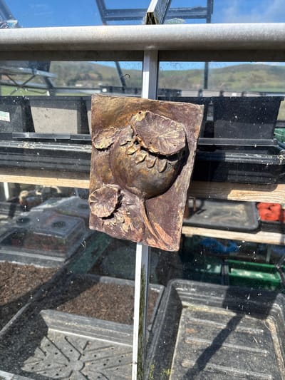The image shows a roughly rectangular, brown and gold-toned, cast sculpture featuring a floral design, lying on a glass surface. The sculpture appears weathered and slightly damaged.

The glass surface is part of a structure, likely a greenhouse or conservatory, as evidenced by the presence of metal framing and multiple panes of glass. Inside the structure, there are stacked black plastic trays and containers, some filled with dark soil.

The location appears to be outdoors, suggested by the view through the glass showing glimpses of a blue sky and distant green landscape. The reflection in the glass indicates a bright, sunny day. Given the request for the location to be Shrewsbury, United Kingdom, the structure can be inferred to be in an outdoor location within the city.