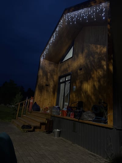 The image shows the exterior of a modern wooden house at night, with string lights illuminating the eaves. The house has a sloped roof and a large window. The foreground is paved with interlocking bricks, and to the left, a wooden staircase leads up to a porch. Various items are scattered on the porch, including bags, containers, chairs, and rolled-up mats. The scene is set against a dark blue night sky and a backdrop of silhouetted trees. The location is Doma 1096 Km, Russia.