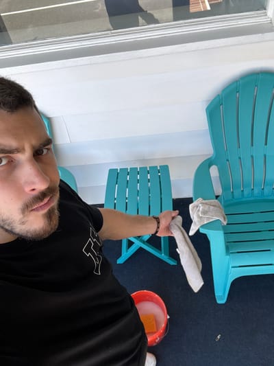 The image, oriented vertically, depicts a man and outdoor furniture on what appears to be a patio or deck. The man, seen from the chest up, has dark hair, a beard, and is looking directly at the camera with a neutral expression. He is wearing a black t-shirt with white outlines of letters, possibly "LA" or similar, visible on his left shoulder/sleeve. His right arm is extended, holding a white towel or rag, suggesting he is in the midst of a cleaning activity. On his wrist, he wears a dark, beaded bracelet.
In the background, there are two turquoise plastic Adirondack-style chairs and a small matching side table. One white towel/rag rests on the arm of the closer large chair. An orange bucket filled with soapy water and a yellow sponge is visible on the dark blue outdoor carpet to the right. The flooring is a dark blue, textured outdoor carpet or rug. To the left, a white wooden wall or siding with vertical panels is visible, along with a window frame reflecting some brick.
The scene is brightly lit, indicative of daytime. The overall impression is that of someone cleaning outdoor furniture on a porch or patio.