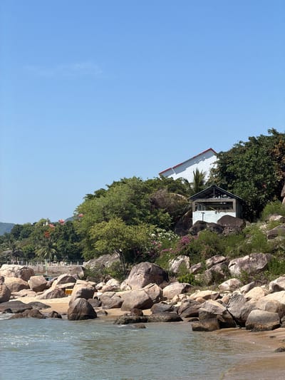 The image shows a tranquil scene of a rocky coastline with lush green trees and vegetation. A small, white building with a red-tiled roof is perched on the hillside, partially obscured by foliage. Next to it is a shaded pavilion with an open design. The coastline is characterized by large, rough-hewn rocks, with some smaller, purplish flowers blooming amongst them. The sea is calm and reflective, with gentle ripples disturbing its surface. In the distance, more greenery and a glimpse of houses with orange roofs are visible, suggesting a populated area. The sky is a clear, vibrant blue, indicating a bright, sunny day. The image appears to be taken from a boat or a low vantage point near the water, looking up towards the shore. The overall mood is peaceful and scenic. There is no visible text in the image.