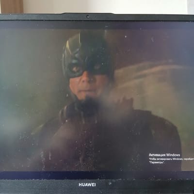 The image shows a laptop screen displaying a person wearing a dark costume with a mask that covers their eyes and the upper part of their face. The background is dark and appears to be blurred or out of focus. In the lower right corner of the screen, there is a Windows activation prompt in Russian, which translates to "Windows Activation: To activate Windows, go to 'Settings'." The text is white and appears over the dark background. The bottom of the laptop bezel is visible with the "HUAWEI" logo. The screen itself has some dust and smudges. The location of Nizhneangarsk, Russia is not depicted in this image, as it shows an indoor scene of a laptop.