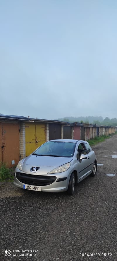 A silver Peugeot 207 hatchback is parked on a gravel road in front of a row of garages in Maardu, Estonia. The garages have variously colored doors, including brown, yellow, and dark gray. The sky is overcast and foggy. The car's license plate reads "202 MGF" and has "EST" in a blue and yellow rectangle, indicating it is registered in Estonia. The lower right corner of the image displays a timestamp: "2026/6/29 05:52". The bottom left corner shows text from the camera manufacturer: "REDMI NOTE 9 PRO AI QUAD CAMERA".