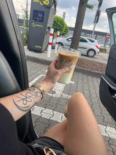 This is a close-up, vertical, eye-level shot taken from inside a car. The focus is on a person's arm and hand holding a clear plastic cup of iced coffee. The person has manicured nails and a large, dark tattoo on their forearm, along with a gold bracelet.

In the background, outside the car, an electric vehicle charging station is visible. The charging station is dark gray with yellow accents and displays various symbols and text, including "EWE" and "e-mobility". Red and white striped bollards are positioned in front of the charging station. A white car is parked nearby, and further in the background, a building with a grid-like facade is visible. A traffic light with a red circular sign is also present.

The scene appears to be outdoors, likely during the daytime, with overcast skies. The paving stones on the ground suggest an urban or parking lot environment. The overall mood is casual, with the individual enjoying a drink. The setting seems to be a public space, possibly a parking lot at a commercial area where electric vehicles are charged.