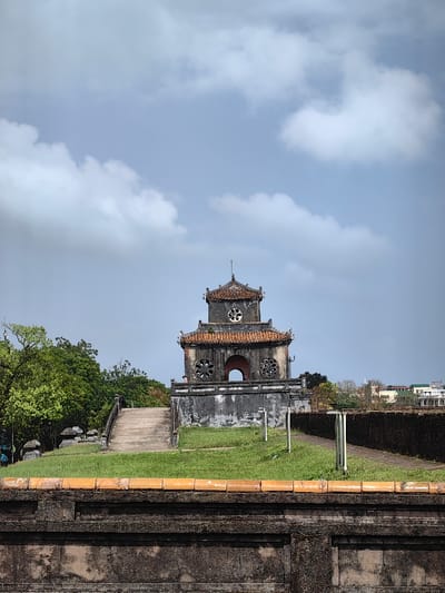 An exterior, eye-level photograph depicts the Hiển Lâm Các (Pavilion of Splendid Caprice) within the Imperial City, Phú Xuân Ward, Huế, Vietnam, under a partly cloudy sky. The central subject is a two-tiered, weathered stone and brick pavilion, characterized by its traditional tiered roof with reddish-brown tiles and dark grey, discolored walls. The lower tier features a central arched opening flanked by two circular, patterned windows. The upper tier displays a single circular, patterned window centrally placed. A small, indistinct human figure is partially visible on the lower tier to the left of the central archway.

In the immediate foreground, a dark grey stone wall with an orange-tiled coping runs horizontally across the bottom of the frame. Behind this wall, a strip of green grass precedes a concrete pathway ascending from the left towards the pavilion. To the right of the grass, a dirt path runs alongside the pavilion, marked by two light-colored vertical posts. Dense green trees and foliage are visible to the left of the pavilion. To the right, a dark stone wall or fortification extends into the midground, beyond which distant urban buildings with light-colored facades and various roof styles are discernible against the horizon. The sky is a light blue, containing scattered white and grey cumulus clouds.