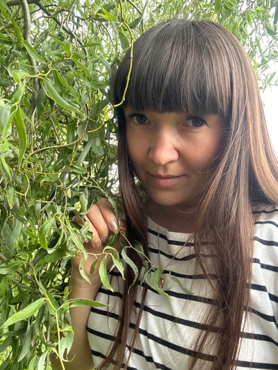 An outdoor, close-up, medium-shot photograph shows a woman with long, brown hair and bangs, wearing a black and white striped shirt. She is positioned to the right of the frame, with her face turned slightly to the left, looking towards the camera. Her left hand is raised and touching some foliage, which also covers the left side of the image. The foliage is green and appears to be from a tree with long, thin leaves. The woman is wearing a thin gold necklace with a heart-shaped pendant. The background is blurred, suggesting an outdoor setting, possibly a park or garden in the city of Sredneye Kechevo, Russia. The lighting is natural and appears to be daytime.