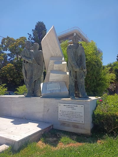 The image depicts a monument in Chalkida, Greece, honoring the National Resistance. The monument features three bronze figures on a marble base, with a large abstract marble structure behind them. Two figures stand together on the left, their arms linked, and a single figure stands to the right, appearing to be manacled. A plaque on the base is inscribed with Greek text, stating "Valor and Courage, Freedom - Kalos" and "The Monument of the National Resistance was erected in 1985 on the initiative of the P.E.D.E.A. Chalkidas and with the symbolic contribution of the prefecture. - Gefthy Georgios Megkoulas." The monument is situated outdoors, with a clear blue sky, lush green trees, and a modern building visible in the background. The lighting suggests it is a sunny day.