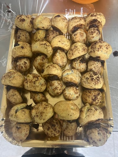 A tray of chocolate-filled cream puffs is presented on a golden tray. The cream puffs are small, round, and have a golden-brown exterior, speckled with dark brown chocolate pieces. Some of the cream puffs are split open, revealing a rich, dark chocolate filling. The tray is placed on a metallic surface, possibly a countertop. The background shows a transparent plastic wrapper and some blurred orange object. The setting appears to be a commercial kitchen or bakery. There are no people visible in the image. The text on the plastic wrapper suggests a connection to Vallo della Lucania, Italy, possibly indicating the origin or sale of these pastries.