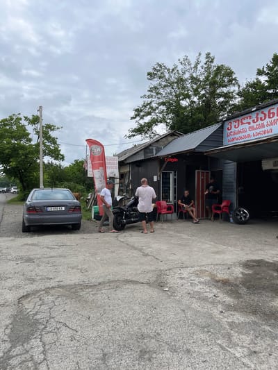 This image captures a street-level view in a rural or semi-urban setting, possibly in Georgia, as indicated by the Georgian text on the signs. The scene appears to be outside a small business, likely a mechanic's shop or tire service, given the presence of a motorcycle and a car.
Several people are present. In the foreground, a man in a light-colored shirt and dark shorts is standing near a motorcycle. Further back, another man in a striped shirt is seated on a chair outside the shop. Inside the shop, a few individuals are visible, one appearing to be working. Two women are seated on chairs in the background.
The setting is outdoors on a cloudy day, suggesting overcast weather. The ground is paved with concrete, showing some wear and tear. The building itself seems to be constructed with corrugated metal and has a sloped, metal roof. A large red banner with Georgian text and a logo is prominently displayed. Another sign above the shop entrance also features Georgian writing. The car parked on the right side of the frame has a license plate that reads "LU-698-KA".
The overall impression is one of casual activity, possibly a break or waiting period at a local service establishment. The presence of people and the ongoing business activity suggest a normal day in this location.