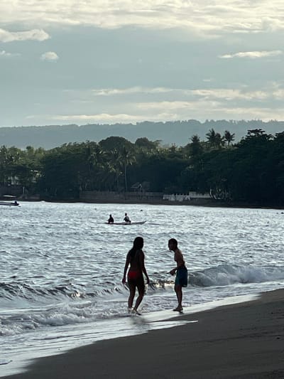 The image captures a serene beach scene in Dauin, Philippines, at dusk. In the foreground, two figures, a man and a woman, stand near the water's edge. The woman, silhouetted against the light, wears a red bikini and faces away from the camera, her body angled towards the man. The man, also silhouetted, wears blue shorts and faces the woman, appearing to be engaged in conversation or interaction with her. The ocean is calm with gentle waves lapping at the shore, reflecting the soft light of the setting sun.
Further out in the water, a long, narrow boat carries two figures, possibly fishermen, paddling through the calm sea. In the background, a dense line of lush green trees and palm trees forms a natural barrier, with a few buildings and structures visible amongst them, suggesting a coastal village or resort. The sky above is a mix of soft blues and whites, with scattered clouds indicating the transition from day to night. The overall atmosphere is peaceful and tranquil, characteristic of a tropical evening.