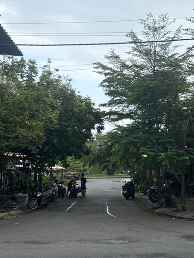 The image depicts a daytime scene on a paved asphalt street in Ngũ Hành Sơn Ward, Vietnam, under an overcast sky. The street surface exhibits two distinct, parallel, curved white marks. Dense green foliage and trees line both sides of the road, obscuring portions of adjacent structures. On the left side of the street, a row of approximately five parked motorcycles is visible along a raised curb. Two young males are present near these motorcycles; one, wearing a red shirt, is leaning towards a motorcycle, while the other, in a dark shirt and shorts, stands upright beside him. A building with a corrugated roof is partially visible above the trees on the left. On the right side of the street, a shaded area beneath trees reveals additional parked motorcycles and at least two partially visible individuals, one of whom appears to be seated. In the distant background, a light-colored pickup truck is partially visible amongst the trees. Multiple black power lines traverse the sky overhead.