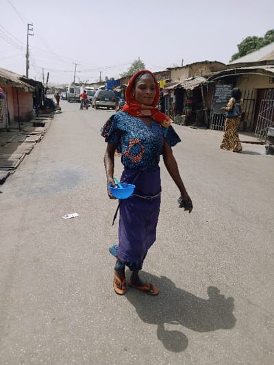 The scene is a daytime street in Masvingo, Zimbabwe, illuminated by bright sunlight casting distinct shadows. A single adult woman stands centrally on a grey asphalt road, positioned slightly to her right and looking towards the viewer. She wears a red and white patterned headscarf, a short-sleeved blue top with an orange and blue pattern, a dark blue wrap skirt, and tan flip-flops with orange straps. Her left hand holds a bright blue plastic bowl containing a small spoon. Her right hand is lowered, grasping a dark, indistinct object. On the road surface near her feet, several small white and pink paper scraps are visible. In the mid-ground, the street continues, revealing a white van, a dark SUV, and a person on a red motorcycle moving away from the viewer. On the right side of the street, another woman in a yellow and black patterned dress stands facing away from the camera, near a building with a large dark chalkboard displaying white text. Buildings with corrugated metal roofs and visible open-fronted shops or stalls line both sides of the street. Utility poles with multiple power lines are prominent on the left side of the street. The sky appears light and hazy.