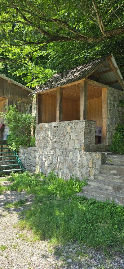This media file depicts an outdoor scene with a rustic, stone and wood structure. The structure appears to be a small building or shelter with a covered roof supported by wooden beams. The walls are constructed from rough-hewn stones, some of which are quite large, interspersed with smaller stones and mortar. The interior of the structure is lined with wooden planks. There's an opening with a floral fabric visible inside, suggesting it might be a small room or alcove.

Adjacent to the structure, there are stone steps leading upwards. The area surrounding the building is lush with green grass and trees, indicating a natural or park-like setting. The lighting suggests it's daytime, with sunlight dappling through the leaves of the trees, casting shadows on the ground. The overall impression is one of a quaint, perhaps secluded, location, possibly a resting spot or a small dwelling in a natural environment. No people or specific activities are evident in the image. The location is identified as Babugent, Russia, which might imply a mountainous or forested region. There is no visible text in the image.