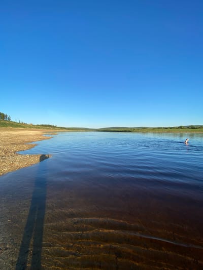 A person is swimming in a wide, dark river under a clear blue sky. The riverbank in the foreground is lined with pebbles and shallow, clear water where a shadow of a person is cast. In the distance, the river stretches towards a tree-lined horizon with rolling hills. The scene is located in the village of Byunty, Russia, a rural area.