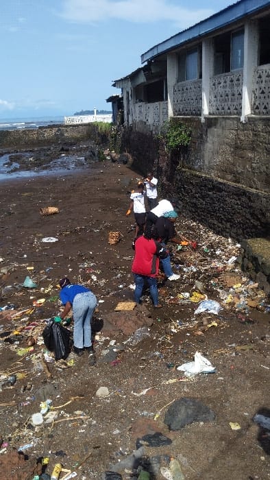 The image shows a group of people cleaning up a beach in Limbe, Cameroon, located at coordinates 4.005 latitude and 9.21 longitude. The beach is covered in debris, including plastic bottles, bags, and other trash. Five people are working together to collect the trash, wearing casual clothes and gloves. The person at the forefront is holding a large black bag and a green tool. The people are standing on a dirt pathway in front of a building with a white wall and a blue roof. The building is located on the edge of the beach. The sky is blue and cloudy.