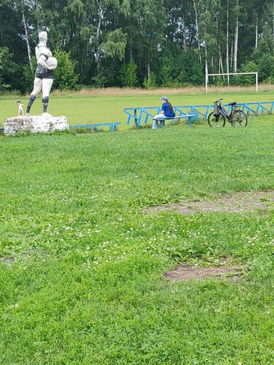 In Drezna, Russia, a statue of a female athlete holding a ball stands on a concrete pedestal in a grassy field. A small dog is visible at the base of the statue. Further in the background, a woman sits on a blue bench next to a bicycle. A soccer goal is situated in the distance behind a line of trees. The environment is an outdoor recreational area, likely a sports field or park, with lush green grass and trees surrounding the scene.