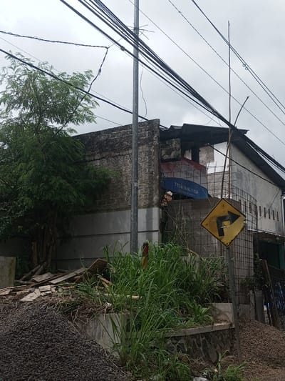 A dilapidated two-story building stands behind overgrown vegetation. A utility pole intersects the view. Power lines crisscross the sky. The building's exterior is weathered, with sections showing brickwork and patched areas. A tilted, faded sign is visible. In the foreground, there's a pile of debris and a street sign with a bent arrow. The sky is overcast.