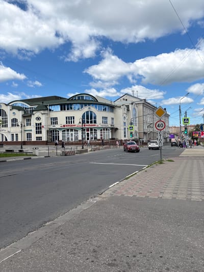 The image shows a street scene in Noginsk, Russia, on a cloudy day. A red car and a white SUV are driving on the road, and pedestrians are walking on the sidewalk. There are several buildings visible, including a bank with the logo "ФОРАБАНК" (ForaBank). Traffic signs indicate a speed limit of 40 and a stop. The sidewalk is paved with bricks, and there are streetlights and traffic lights. The sky is blue with white clouds, and there are power lines visible across the sky.