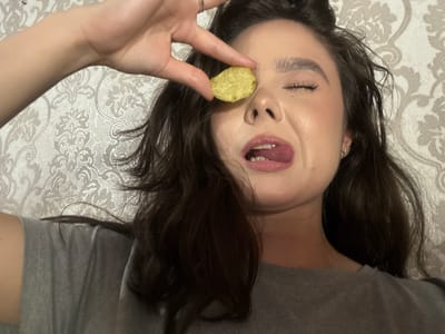 A young woman, with dark, wavy hair and fair skin, is captured in a close-up selfie. She is wearing a grey t-shirt and has her eyes closed, with her tongue sticking out playfully. A single potato chip is held between her index finger and thumb, positioned over her right eye. The background is a textured, light-colored fabric with a damask pattern. No specific location in Zhovte, Ukraine, is identifiable.