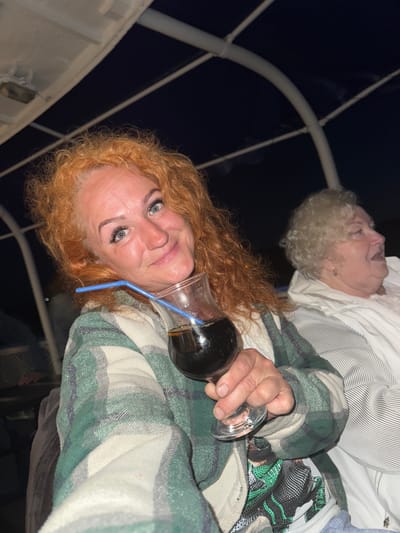A woman with bright orange, curly hair and blue eyes smiles as she holds a glass of dark liquid with a blue straw in it. She is wearing a green and white plaid jacket over a t-shirt with a graphic design. To her right, an older woman with short, curly blonde hair is looking away from the camera. Both women appear to be seated on a boat, with the dark night sky and a white canopy structure visible in the background. The woman's left hand is holding the glass, with her fingers curled around the stem. The image is taken from a low angle, looking up at the women. There is no visible information to place this image specifically in Dulesovo, Russia.