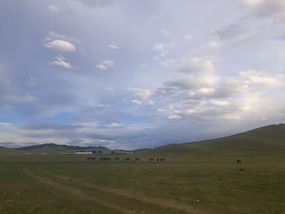 The image presents an expansive grassland environment near Ondersant, Mongolia, under a partly cloudy sky. In the foreground, two parallel dirt tracks traverse the green terrain. Approximately seven dark-colored horses are observed in the mid-ground, clustered together and appearing to graze. A solitary dark-colored horse is visible grazing further to the right in the mid-ground. In the distant left-middle ground, several low-lying structures, including one with a reddish roof and others appearing light-colored, are discernible against the base of distant, rolling hills. A large green hill rises prominently on the right side of the frame. The sky above is a mix of light grey and darker blue-grey clouds, interspersed with brighter white cumulus clouds. No people are visible.