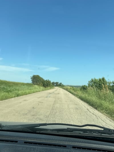 A roadside scene, likely taken from inside a moving vehicle. A concrete road stretches forward, flanked by green grass and low-lying trees on either side. Above the road, a clear blue sky is visible. In the foreground, the vehicle's dashboard and a windshield wiper are present, indicating the viewpoint. The location is suggested as Septemvritsi, Bulgaria.