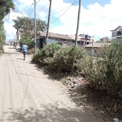 The scene depicts an unpaved dirt road in Ongata Rongai ward, Kenya, under a partly cloudy sky. One adult male, wearing a blue patterned shirt and dark trousers, walks away from the camera on the left side of the road. The road surface shows tire tracks and scattered shadows. Along the right side of the road, dense green bushes are visible, beneath and within which a substantial accumulation of refuse, including plastic bags and bottles, is present. Multiple wooden utility poles with overhead wires line the road. In the background, various buildings are visible, ranging from single-story structures with corrugated iron roofs to multi-story residential buildings, some appearing incomplete, including one with an elevated black water tank. A white vehicle is parked or moving slowly further down the road on the left.