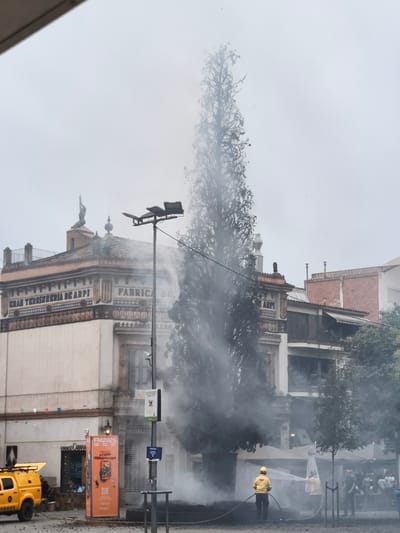 The image depicts a scene in the city of Sant Cugat del Vallès, Spain, specifically in front of the building labeled "GRAN TERRISSERIA DE ARPI" and "FABRICA DE". A tall, slender tree is enveloped in thick smoke, appearing to be on fire or experiencing an event that is producing a significant amount of smoke. Two individuals, likely firefighters based on their yellow uniforms and helmets, are actively engaged in spraying water from hoses towards the base of the tree and the surrounding area. One firefighter is clearly visible in the foreground, directing a stream of water, while another is partially obscured by smoke in the background. A yellow utility vehicle with its hazard lights on is parked on the left side of the frame. In front of the "GRAN TERRISSERIA DE ARPI" building, there is an orange advertising stand for "FM2026". The overall atmosphere suggests an emergency situation involving a fire or a controlled burn being extinguished in a public space.